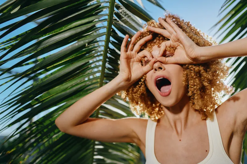 playful woman with curly hair making binoculars gesture under palm leaves, joyful expression, sunny summer day, natural light, outdoor tropical vibe