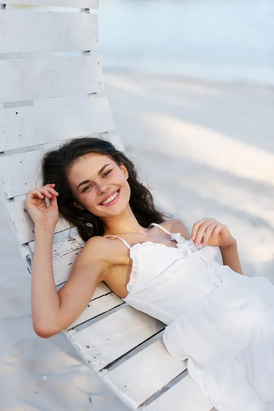 Young woman in a white sundress lounges on a weathered wooden bench by the beach, smiling softly as sunlight highlights her relaxed pose and carefree style.