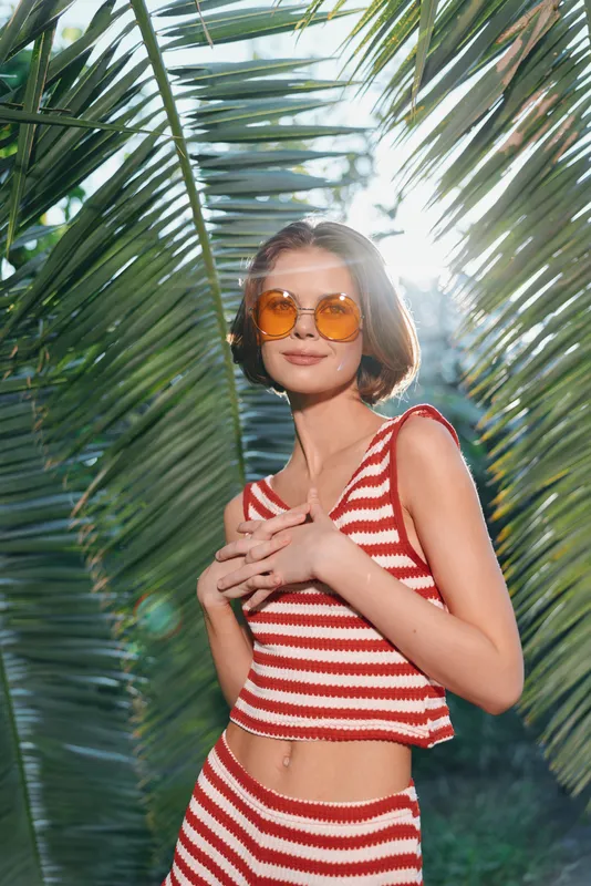 A cheerful model wears a red and white striped bikini top in a tropical setting. She poses with a confident smile amid lush palm leaves, creating a bright sunny summer vibe.