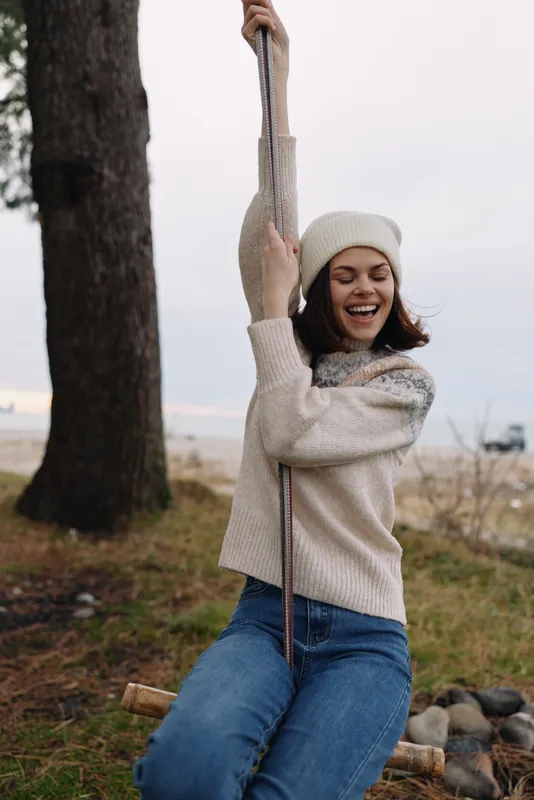 Casual outdoor scene featuring a woman on a rope swing in a park, wearing a knit sweater and beanie, laughing while gripping the rope, with trees and a natural ground setting nearby