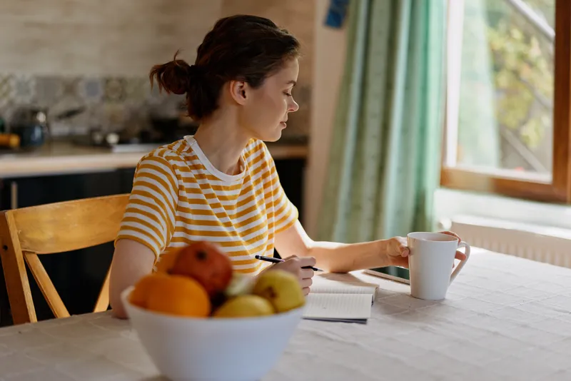 A woman sits at a kitchen table, writing in a notebook as sunlight pours through a window. A fruit bowl and mug complete a calm home moment.