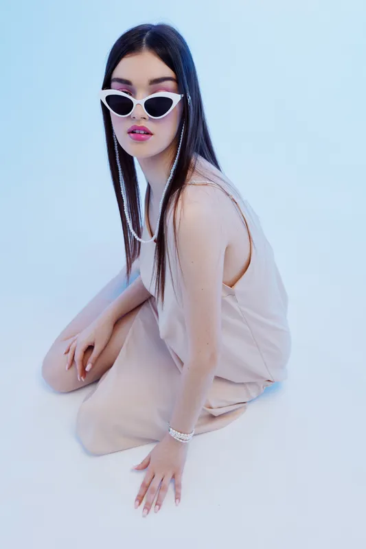 Young woman in beige dress and white sunglasses poses on floor against light blue background. Concept for woman beauty fashion and modern style.