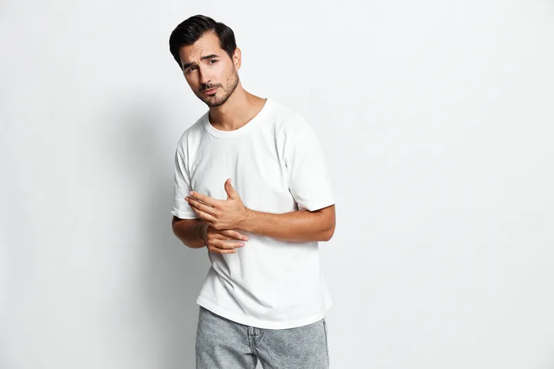 Handsome young man in casual white t-shirt posing thoughtfully with hands together, looking at the camera against a plain light background. Modern lifestyle concept.