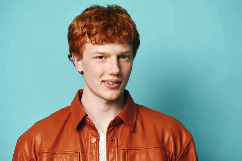 Young man with red hair wearing a brown leather jacket and white shirt posing with smile on blue background in studio. Casual portrait, confident expression and hairstyle.
