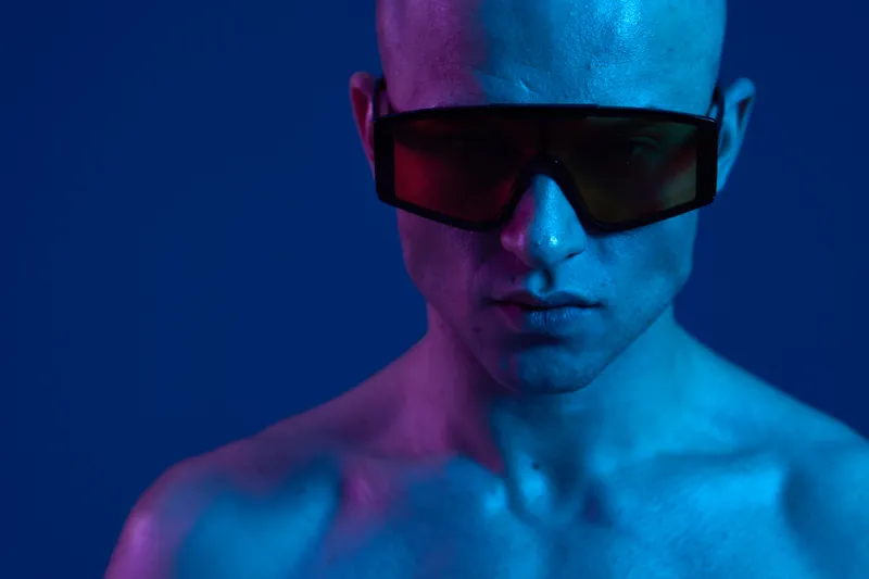 A striking man with shaved head, wearing designer sunglasses in a vivid color light setting, exudes confidence and modern style. Perfect for fashion themes.