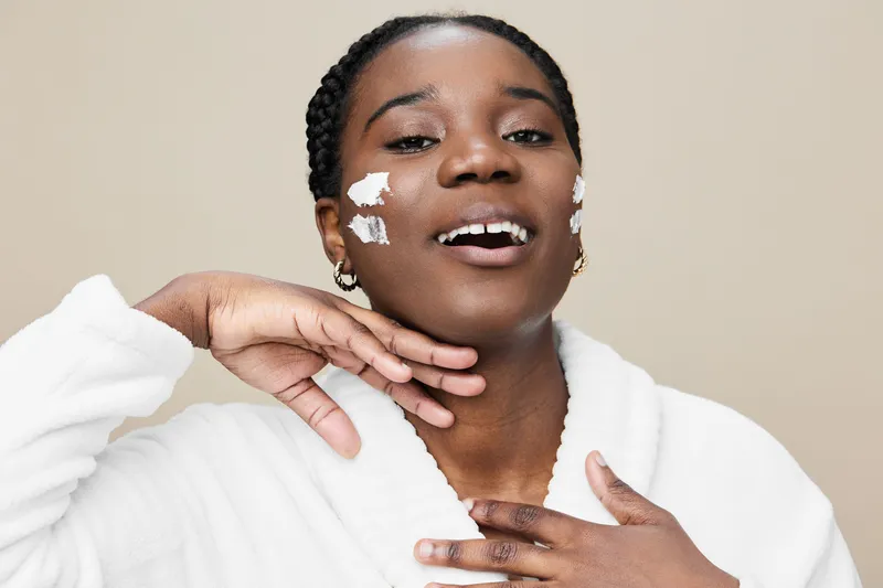 Smiling woman applying skincare product with facial cream on cheeks, wearing a cozy bathrobe against a neutral background, promoting beauty and self care