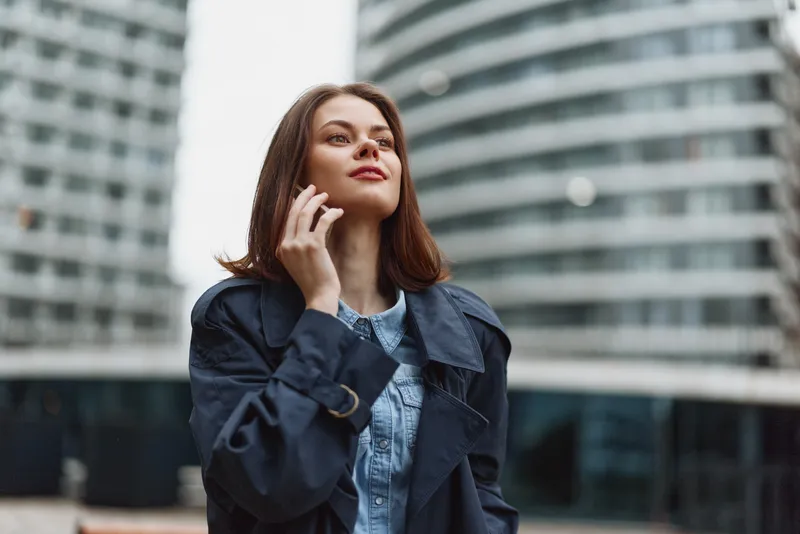 Young woman with brunette hair in a dark trenchcoat looks up as she talks on her phone against a modern city backdrop, exuding confidence and curiosity amid glass towers.