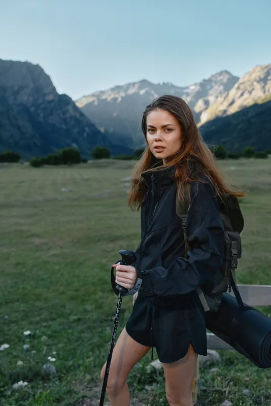 A focused photographer stands in a wide mountain valley, holding a camera and tripod. She wears a dark jacket and backpack, ready to capture dramatic outdoor scenery.