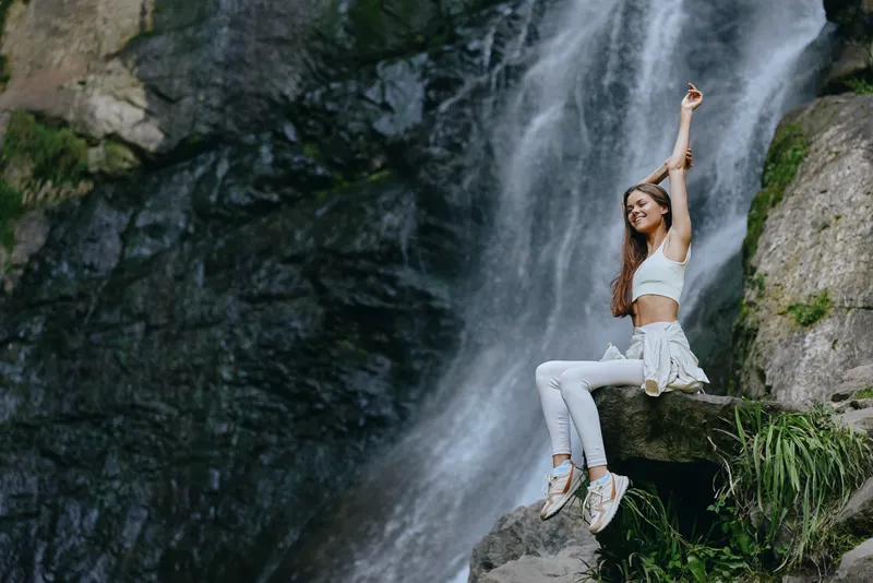 young woman enjoying nature sitting on rock near waterfall, wearing casual white clothes and sneakers in peaceful outdoor environment