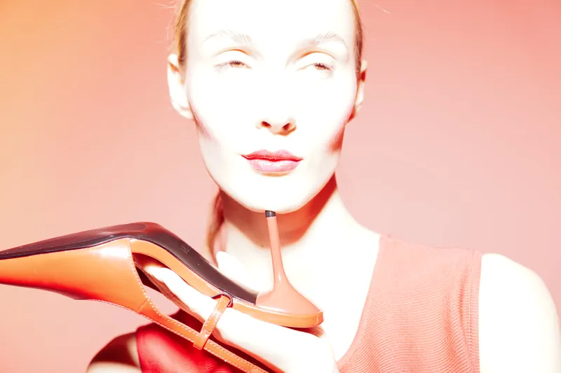 Woman with pale skin holds red high heel shoe near face against orange background. Fashion concept with bright lighting and minimalistic style focusing on footwear and beauty.