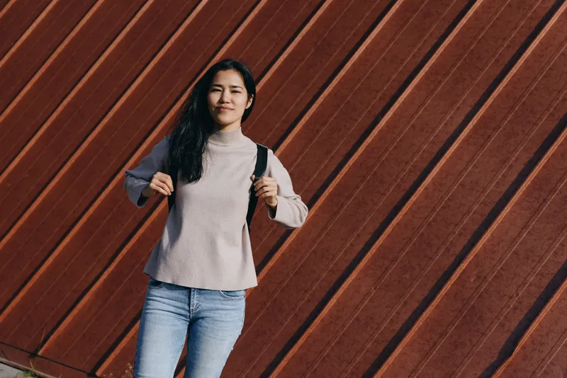 Woman with backpack standing against a diagonal wooden wall in a casual outfit, relaxed pose, confident expression, outdoor urban setting, street style.