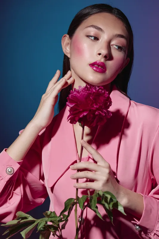 Young woman with slick hair and shiny pink lipstick holds bright flower in delicate pose. Soft studio lighting emphasizes beauty and fashion concept with creative styled makeup.