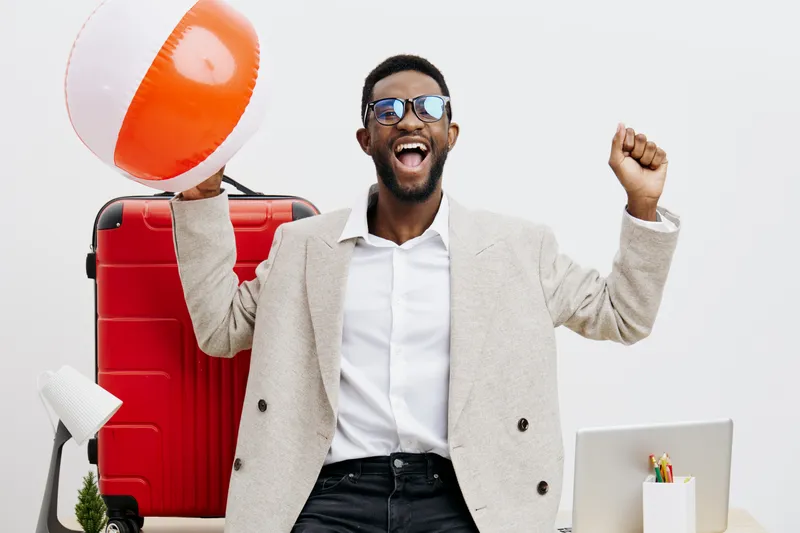 Happy cheerful man with sunglasses celebrating at office, holding inflatable beach ball, with red suitcase and travel accessories, on white background. Vacation, travel, vacation mood, celebration, ex