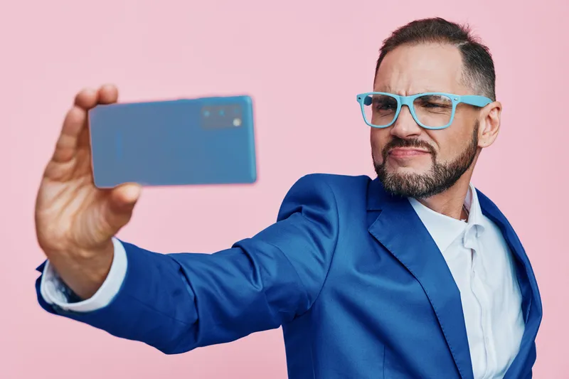 Professional man in a blue suit poses against an isolated pink background, exuding confident energy as he takes a selfie, showcasing bold emotion and modern business style.