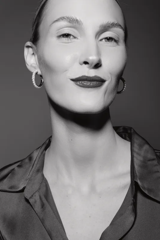 Close-up portrait of a white adult woman with pale skin and earrings, wearing dark clothing and subtle makeup in a studio setting.