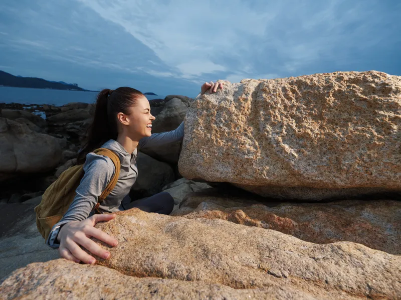 Smiling young woman enjoying outdoor adventure while climbing rocks at the beach, wearing sporty attire and a backpack