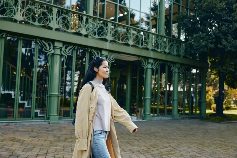 Woman walking outdoors in a beige coat along a glass building facade, casual fashion, daytime urban scene, candid street style and relaxed movement and pose