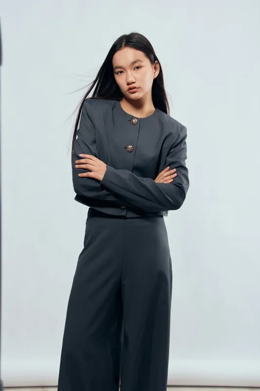 Young woman with long black hair wearing modern dark gray business suit standing with crossed arms looking confident in studio with plain light background and minimalistic style.