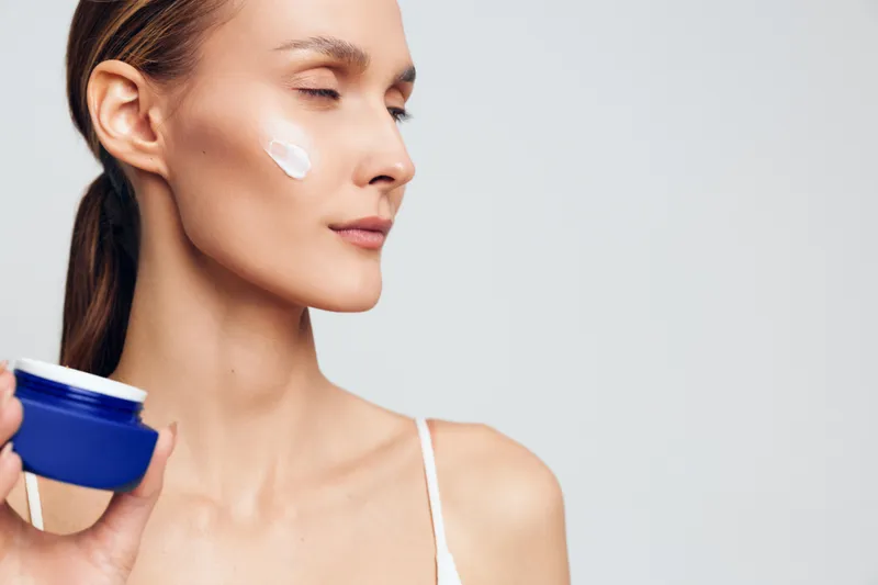 Woman skincare close up face cream application with cotton pad holding blue container calm expression studio white background beauty hygiene routine morning care
