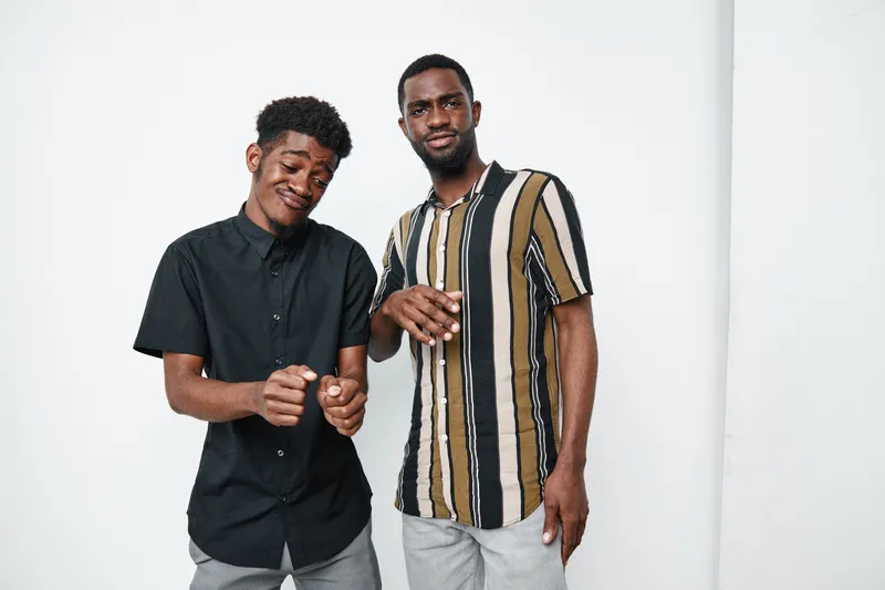 Two young black men with dark skin posing together in casual shirts against white background, smiling and showing relaxed friendly gestures in studio portrait.