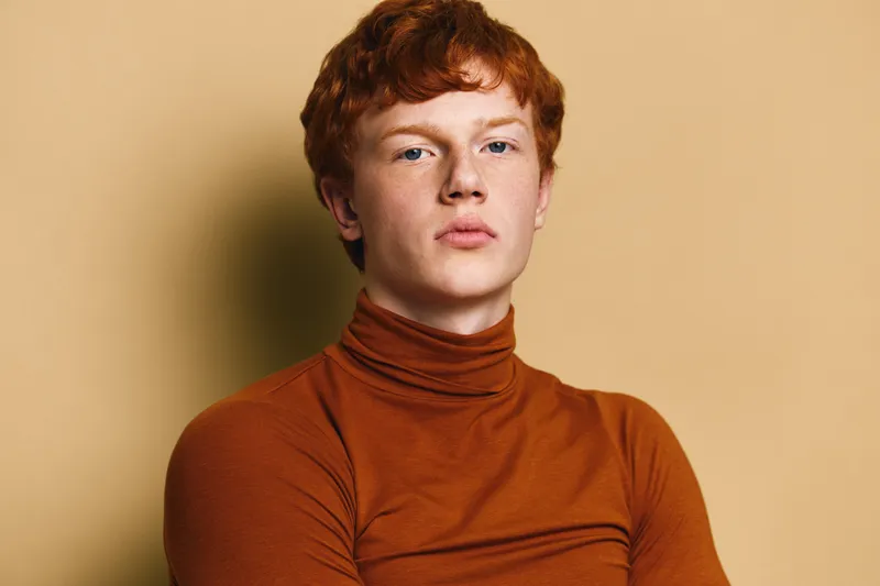 Portrait of a young redhead man wearing a brown turtleneck sweater standing against beige background, looking confident serious natural skin and hair details captured in studio lighting.