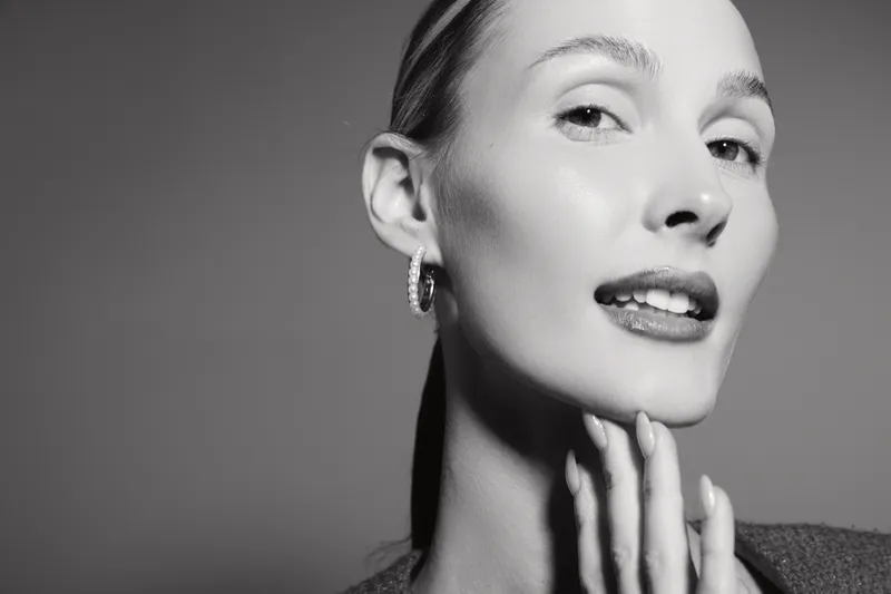 Close-up portrait of a fair skinned white woman with slicked back hair touching her chin, posing confidently against neutral background in black and white.
