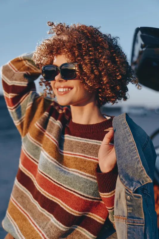 Woman lifestyle, curly hair, sunglasses, smiling young woman in striped sweater and denim jacket outdoors in bright sun, close up by seaside car, summer travel, vacation vibe