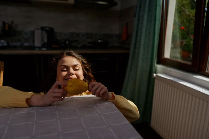 Smiling woman with curly hair sits at a home kitchen table, holding a ripe mango. Close-up on a cozy interior, natural light, relaxed mood, everyday wellness and snack moment.