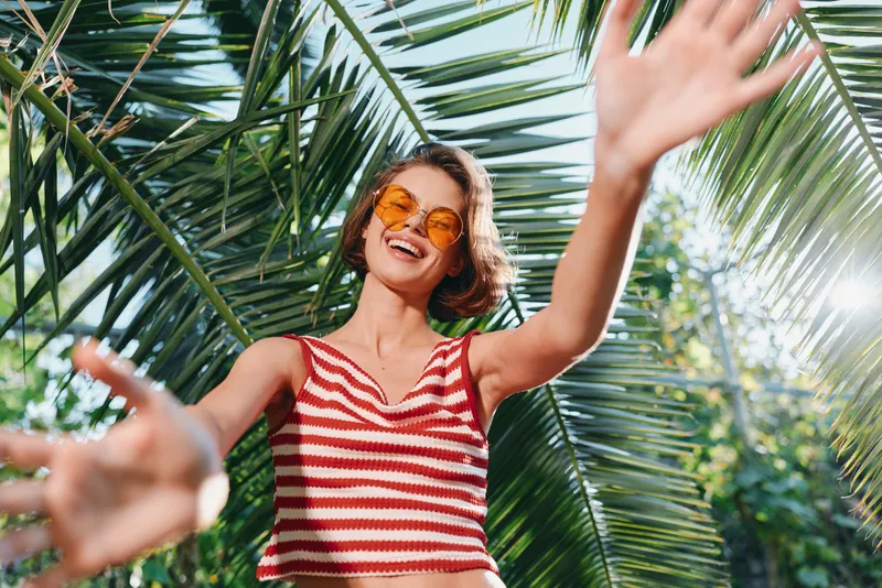A cheerful woman stands outdoors among lush palm leaves, wearing a striped crop top and sunglasses. She smiles broadly, radiating a sunny, energetic street style and positive mood.