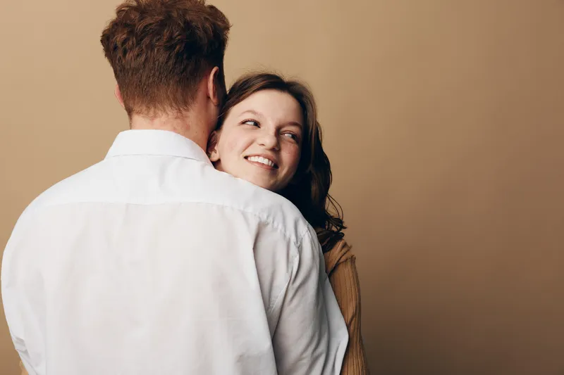 Young couple embraces tenderly with woman smiling and looking over man's shoulder. Intimate moment captured in warm studio with beige background. Concept of love, affection, and connection.