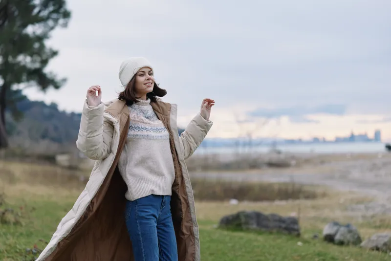 Woman outdoors in a long coat, cozy sweater and beanie, enjoying a breezy day by a lake with open space and distant hills, feeling calm and stylish.