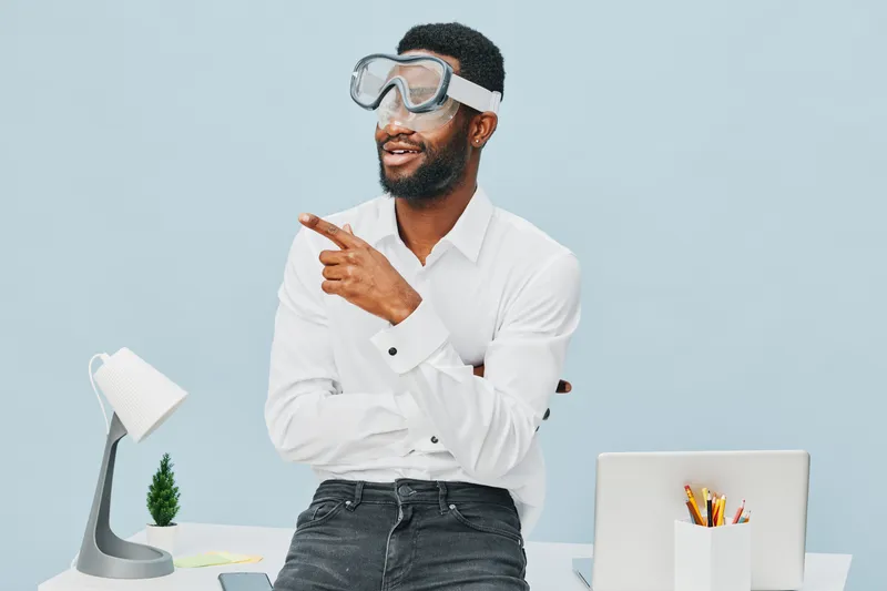 Confident man with beard wearing goggles and white shirt pointing upwards indoors with modern office desk setup, creative workspace, and a laptop, emphasizing innovation and smart ideas. Office lifest