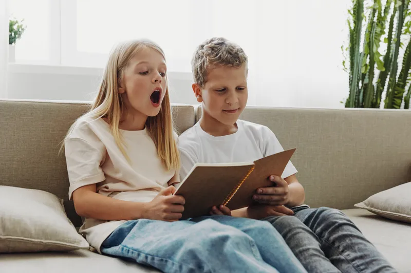 Two children sitting on sofa reading a book together, girl showing surprise, boy focused on the pages. Cozy home environment, learning and curiosity concept.