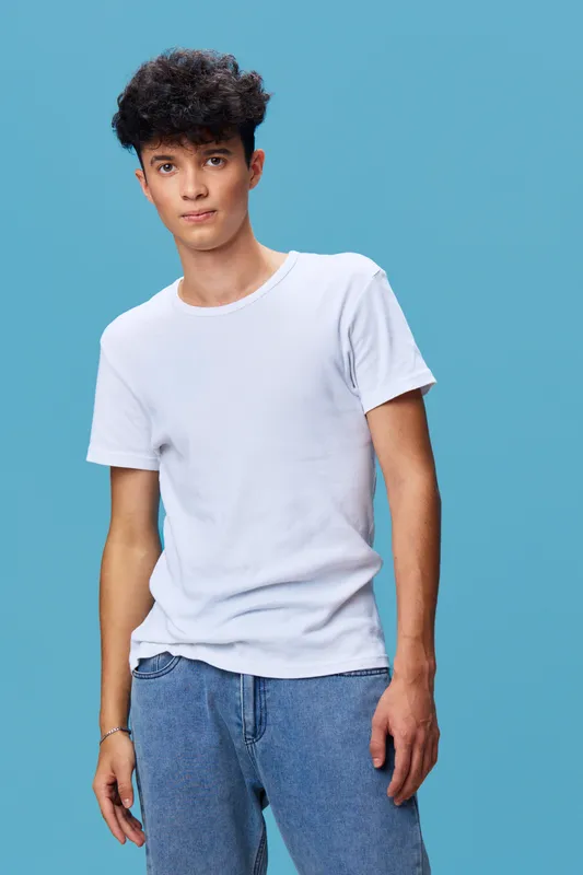 young man wearing white t-shirt and blue jeans standing against blue background with casual posture and curly black hair, studio portrait with soft lighting and relaxed expression