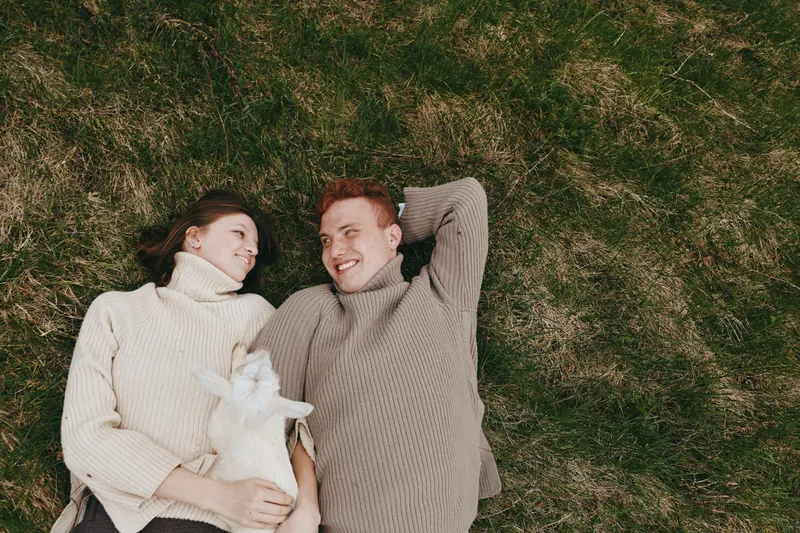 Cheerful couple outdoors, lying on grass in cozy sweaters, sharing a warm smile while a small animal rests nearby, sunlit moment, natural colors, relaxed posture, affectionate mood, casual lifestyle,