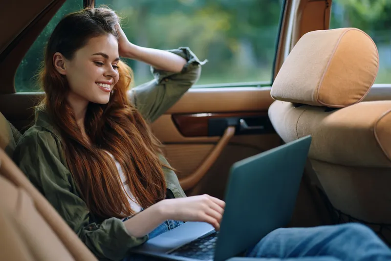 Young woman with long hair relaxing in a vintage car, smiling while working on a laptop in a natural, vibrant setting during summer.