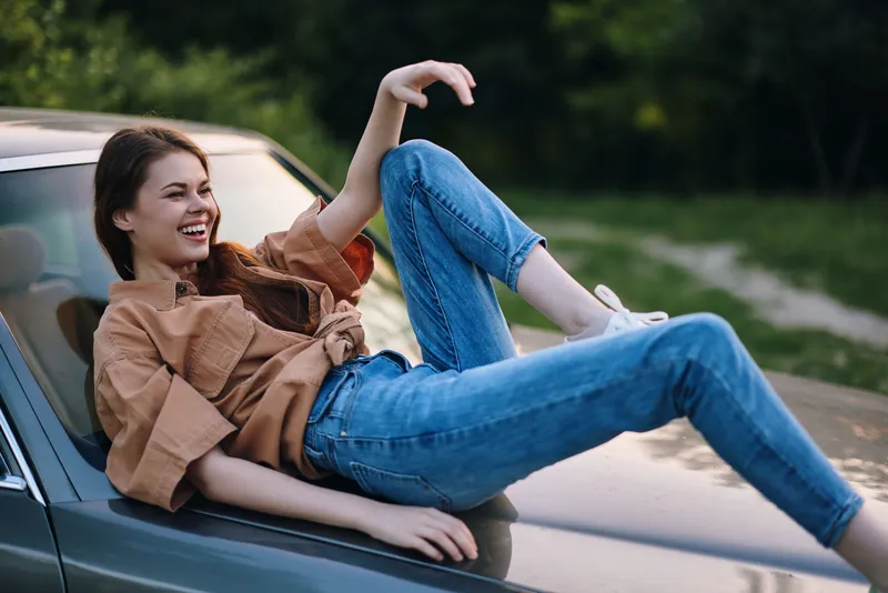 Young woman joyfully lounging on a vintage car, wearing casual denim and a stylish brown top, capturing a carefree summer vibe in a lush green setting.
