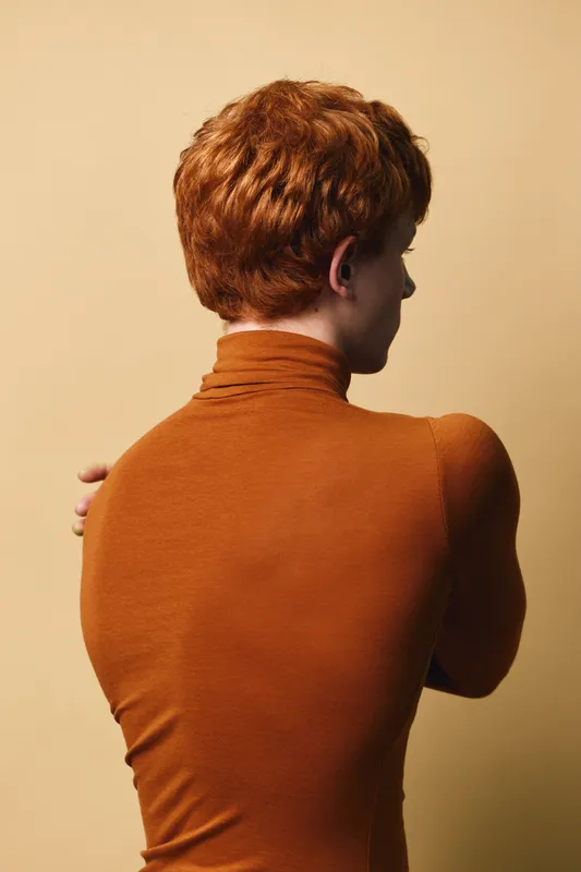 Person with red hair and brown turtleneck sweater facing away against beige background, arms crossed behind back, showing side profile and smooth skin texture in studio lighting.
