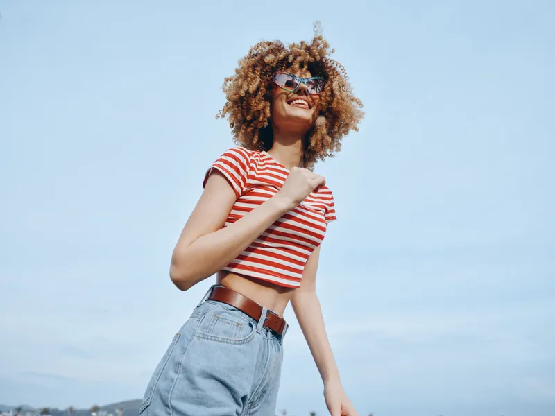 A youthful woman stands outdoors in bright daylight, wearing a red and white striped crop top, denim jeans, and sunglasses, smiling confidently toward the camera.