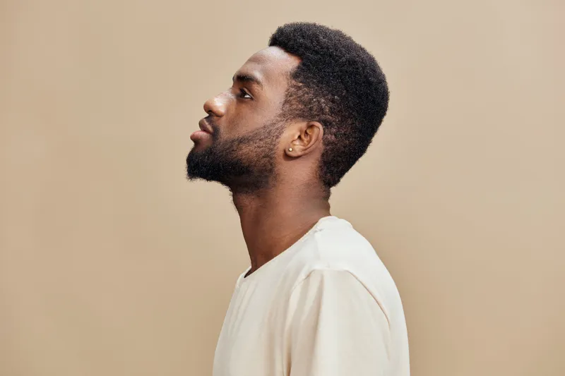 Profile portrait of a young Black man with a short beard, wearing a light shirt against a neutral background, showcasing a contemplative expression and minimalist style