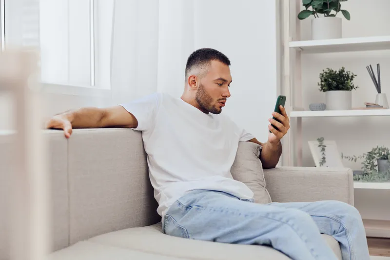 Relaxed man sitting on a sofa, engaging with his smartphone, in a modern and bright living room with minimalistic decor and greenery. Perfect for lifestyle themes in 2025.