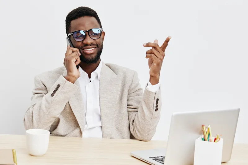 Smiling man with dark skin, glasses, white shirt and beige blazer talking on mobile phone while sitting at a desk with laptop and stationery, studio background. People lifestyle concept