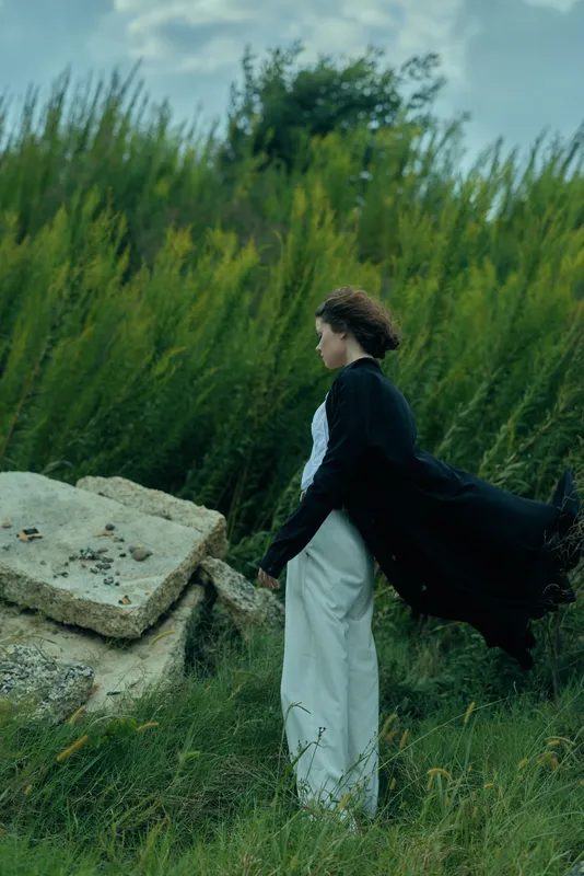 Cape wearing woman outdoors with wind blowing her cloak in a field, fashion mood, mysterious silhouette, graceful movement, nature scenery, serene atmosphere, dramatic sky, cinematic style
