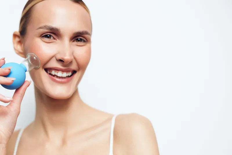 Young woman with blonde hair smiles brightly while using a facial suction device on her cheek against a clean white background.