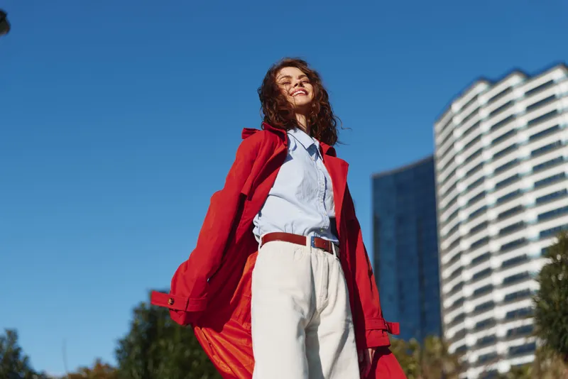Young woman in a bold redcoat stands outdoors under a clear blue sky, white shirt and light trousers, urban buildings behind, exuding confidence and joy.