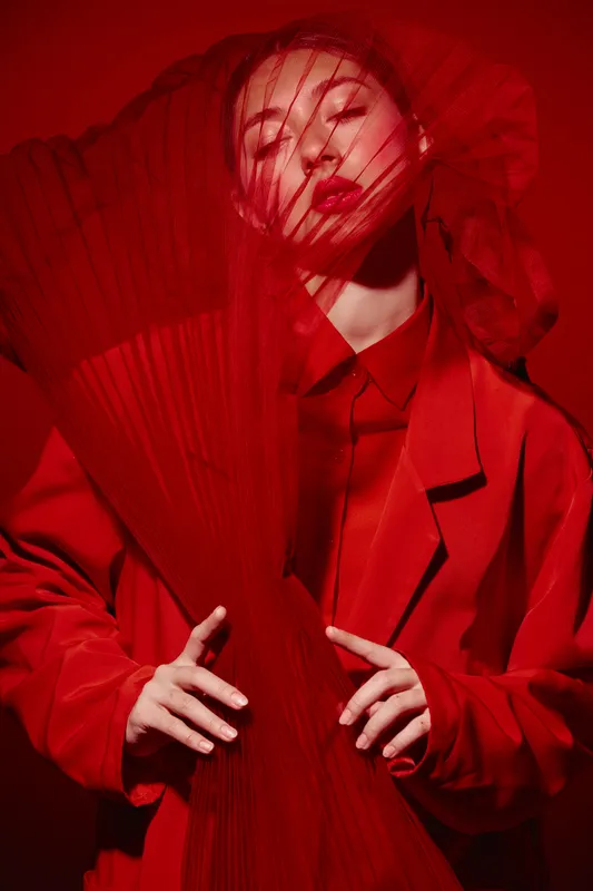 Creative fashion art portrait of a woman wearing red coat and veil. Dramatic lighting highlights vibrant texture and deep hues, capturing stylish artistic beauty concept andavantgarde.