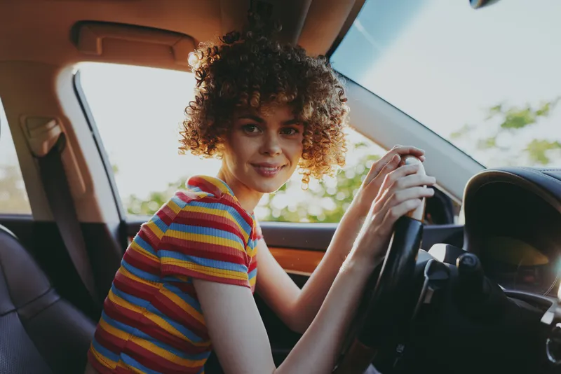 Woman driver sits in car interior, smiling at camera, wearing striped shirt, casual lifestyle moment, warm retro film color tones, daylight through window, candid portrait.