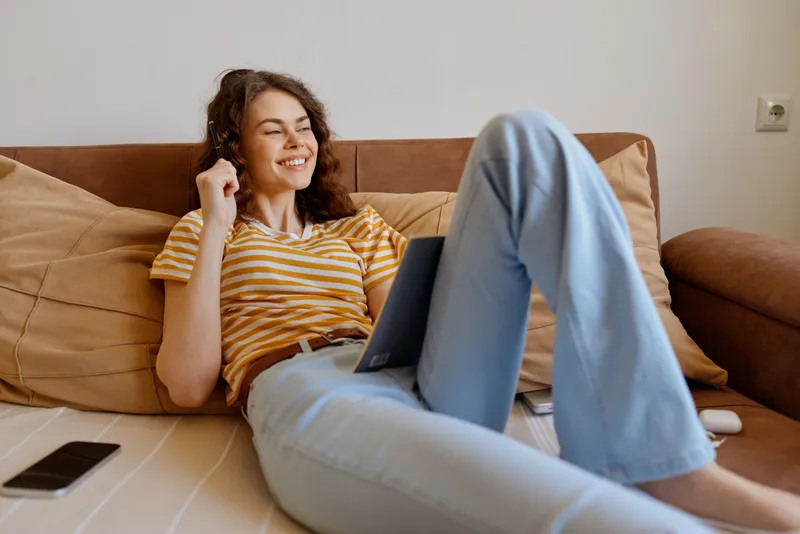 A woman relaxing at home on a couch with a laptop, casual lifestyle scene in a bright, comfortable living room. A moment of everyday joy, emphasizing home life and digital work.