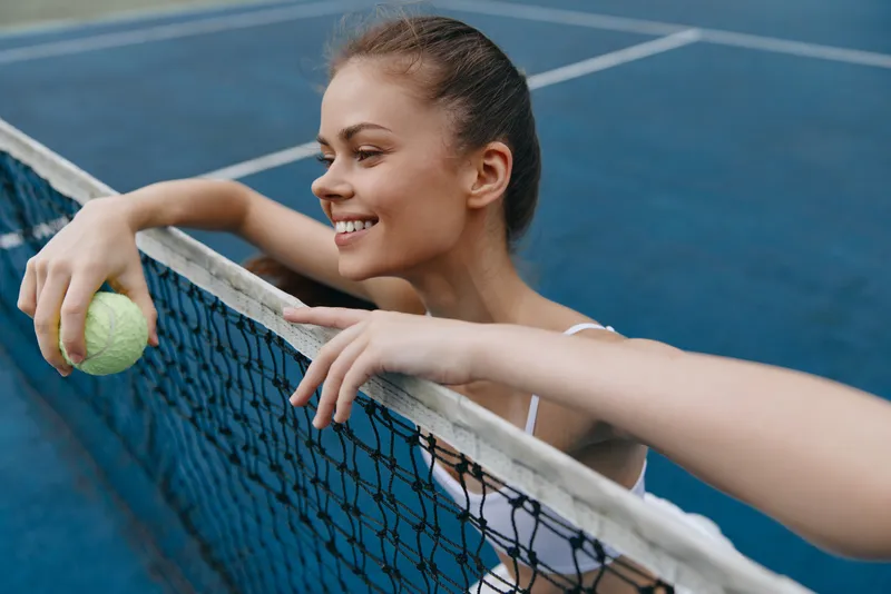 Smiling young woman playing tennis, wearing athletic wear and holding a tennis ball, against a vibrant blue court background