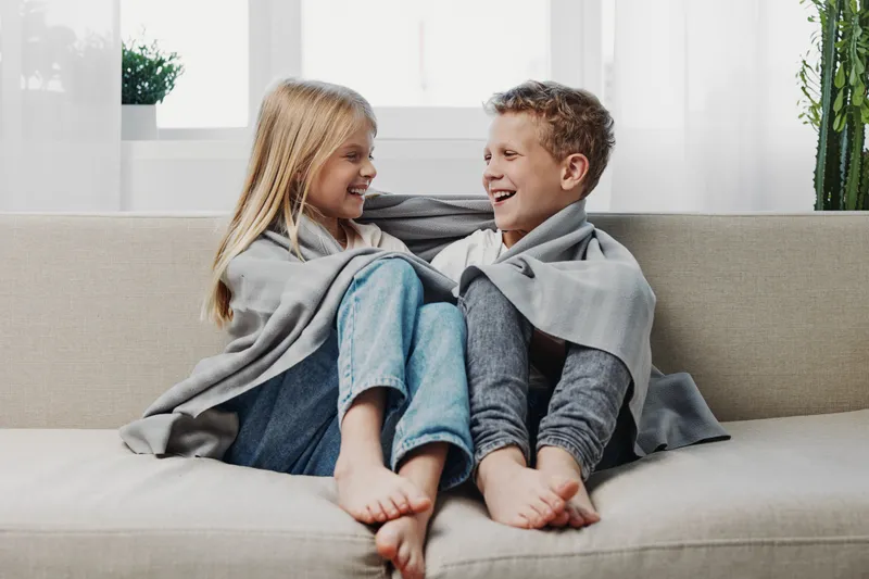 Happy children wrapped in grey blanket sitting barefoot on couch smiling and looking at each other in bright living room.